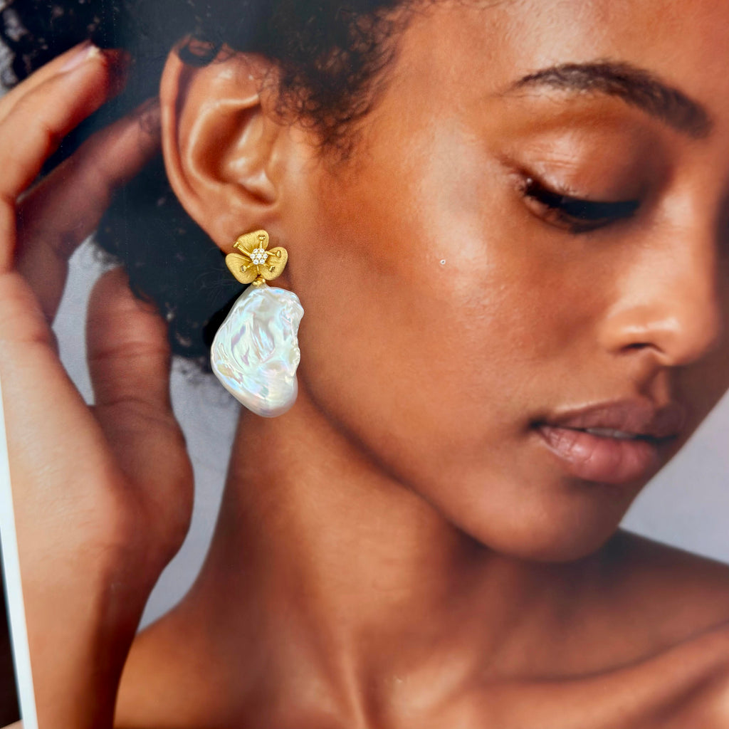 A close-up lifestyle shot of a model wearing a unique drop earring. The earring features a gold flower-shaped stud with tiny clear stones, hanging from a large, irregular white pearl-like drop.