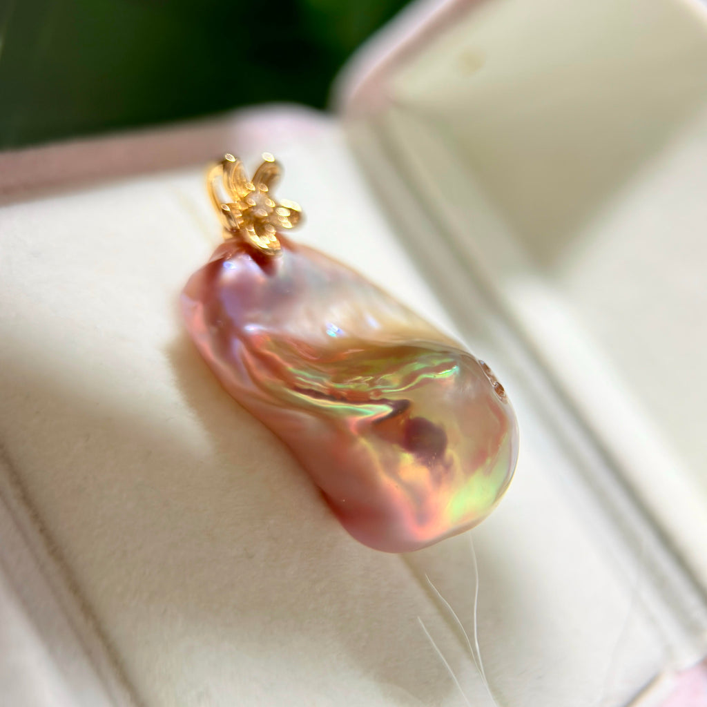 A close-up of a baroque-style freshwater pearl pendant with a gold flower-shaped bail, resting inside a white jewelry box.