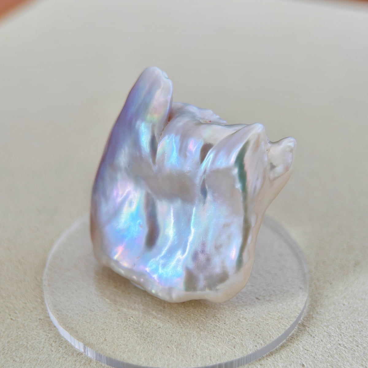 A baroque-style iridescent freshwater pearl displayed on a clear acrylic stand against a neutral beige background.
