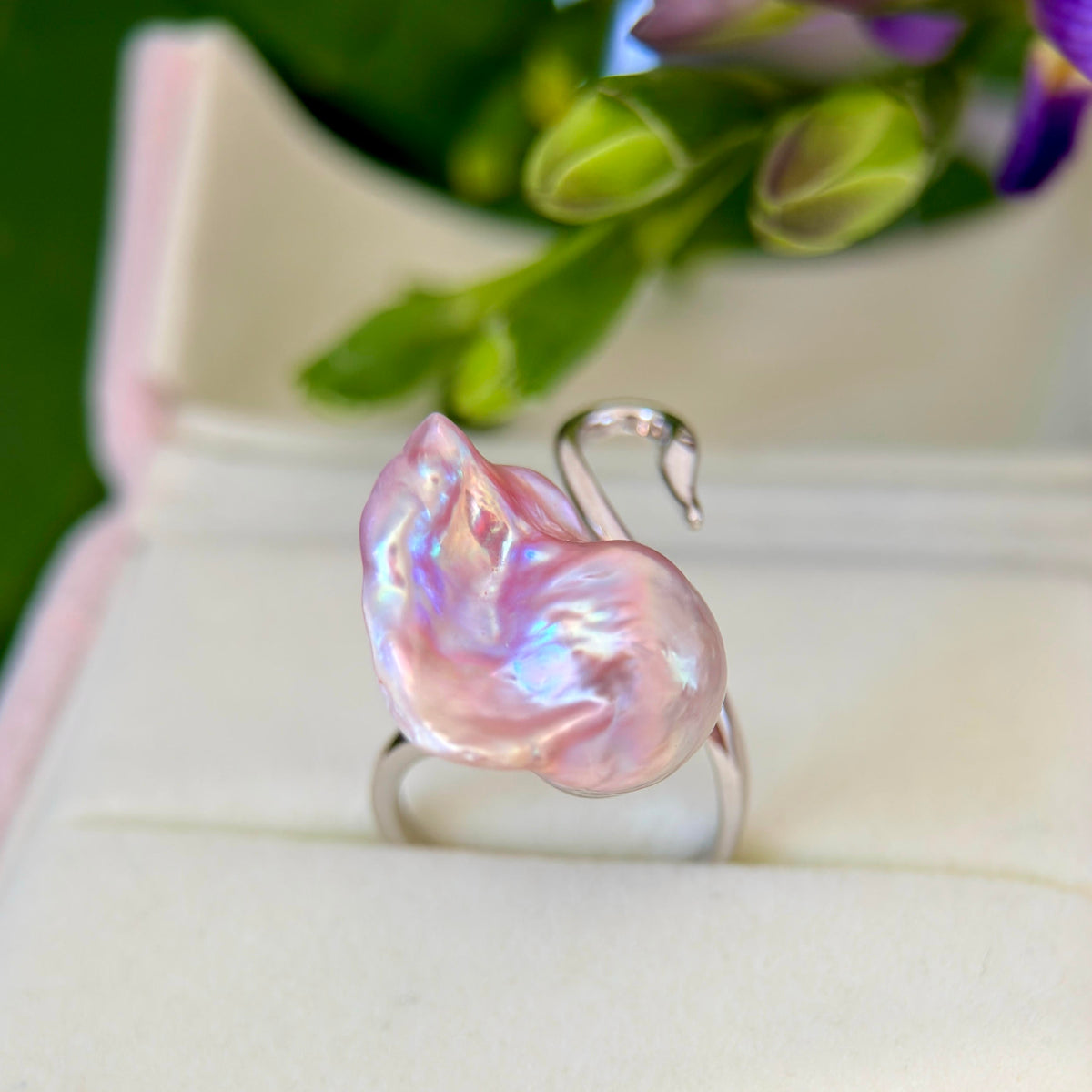 A silver ring featuring a large, irregularly shaped pink baroque pearl with iridescent blue and purple flashes, displayed in a white ring box with blurred green leaves in the background.