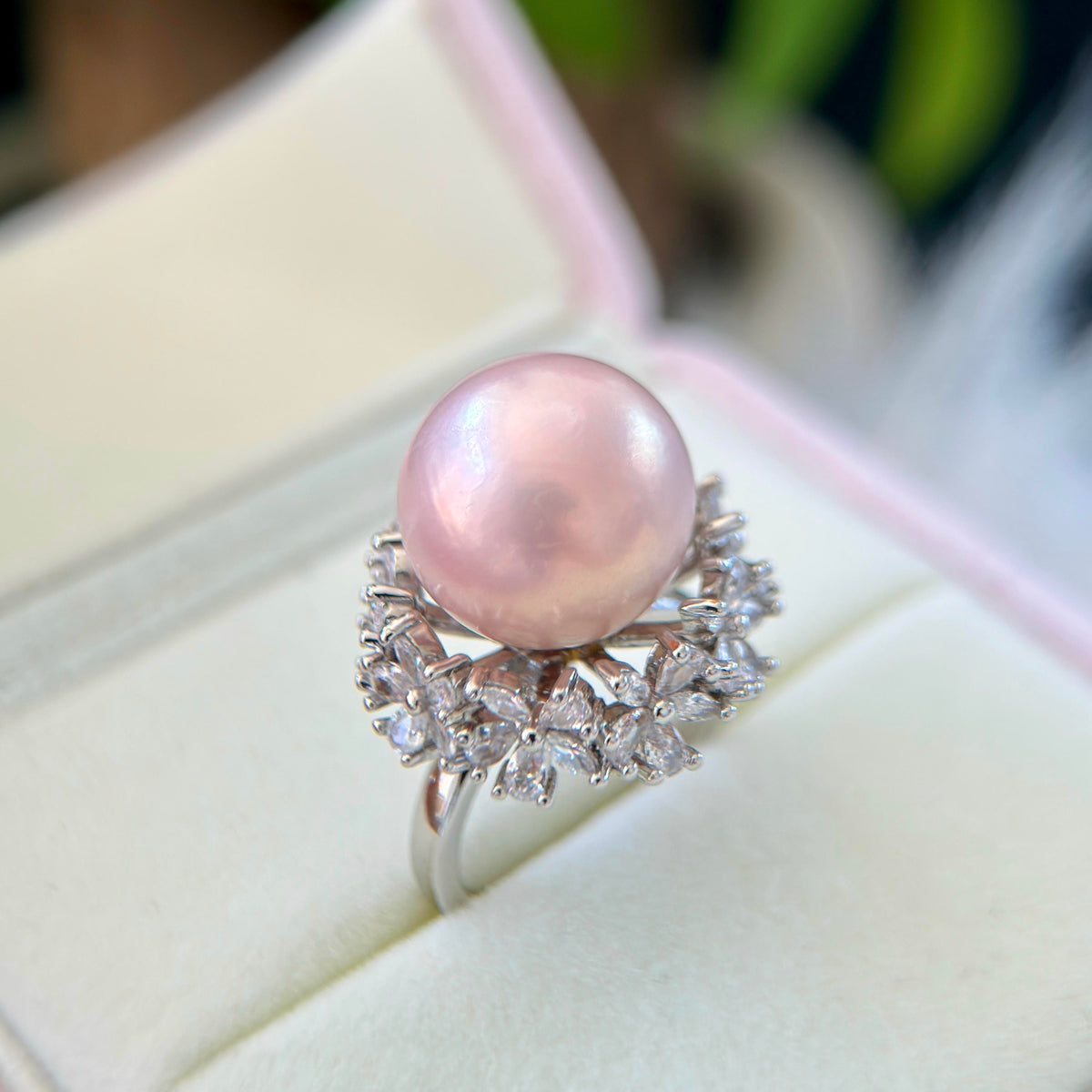 A silver ring featuring a large, lustrous pink pearl surrounded by small clear marquise-cut stones, displayed in a soft pink-lined jewelry box.