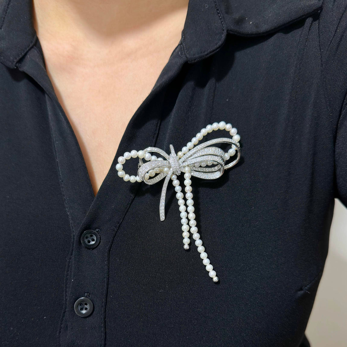 A silver bow-shaped brooch embellished with white faux pearls, pinned to the chest of a black button-up shirt.