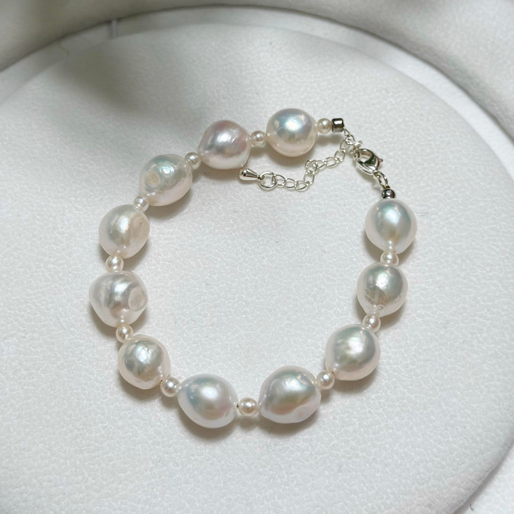 A beaded bracelet made of irregular white baroque pearls and small silver spacer beads, connected by a silver chain with a lobster clasp, displayed on a white textured surface.