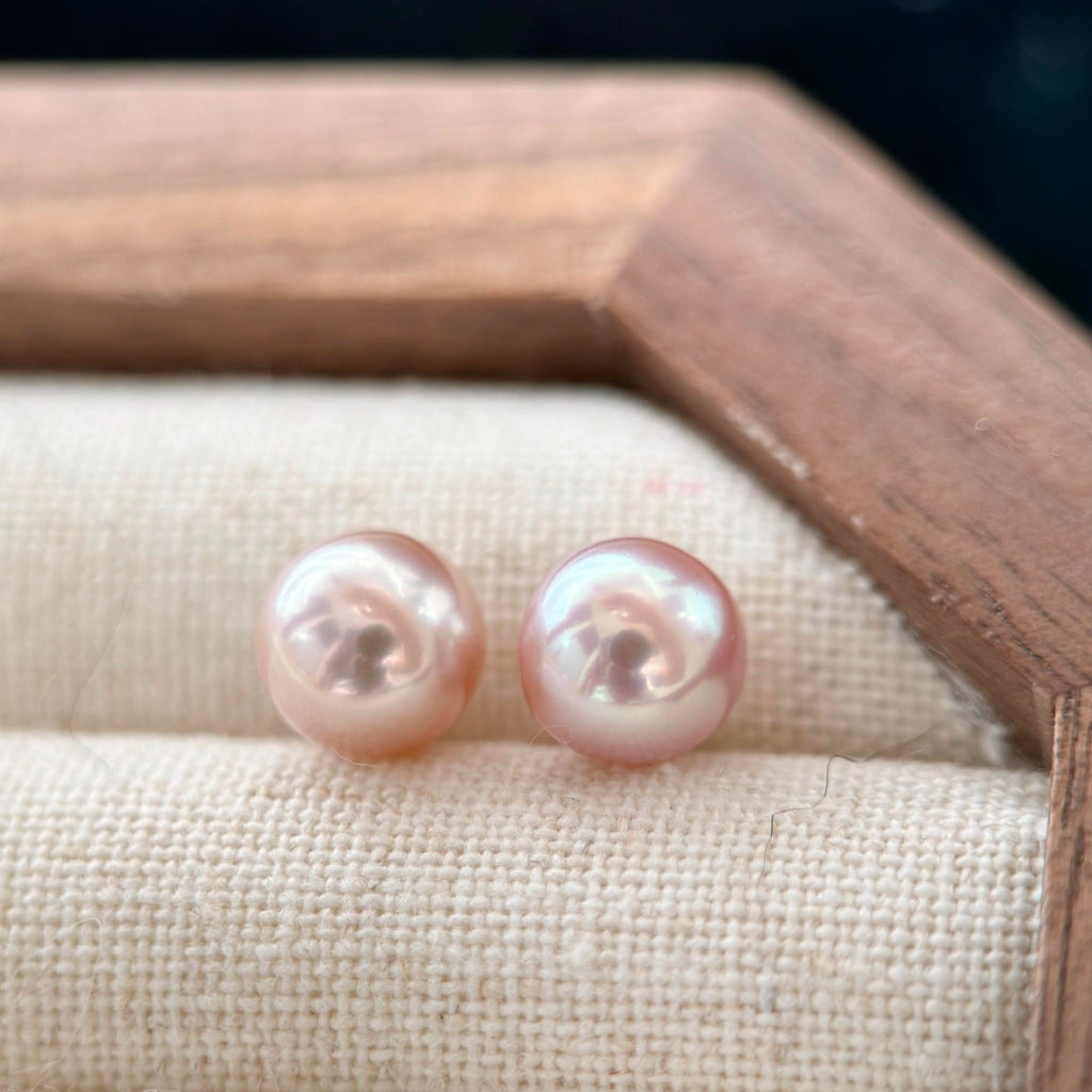 A pair of round freshwater pearl stud earrings with a soft pinkish-gold luster, resting on a textured beige fabric cushion inside a wooden jewelry box.