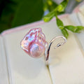 A silver ring featuring a large, irregular pink baroque pearl and a curved silver snake head, displayed on a cream jewelry box with blurred green leaves in the background.