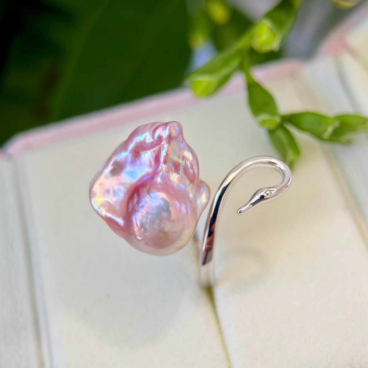 A silver ring featuring a large, irregular pink baroque pearl and a curved silver snake head, displayed on a cream jewelry box with blurred green leaves in the background.