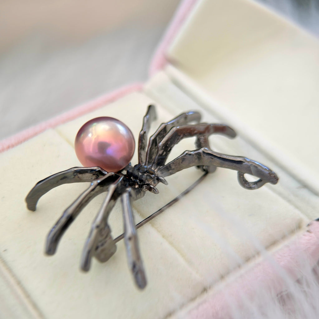 A spider-shaped brooch with a glossy pink pearl and dark metallic legs, displayed on a cream-colored jewelry box.