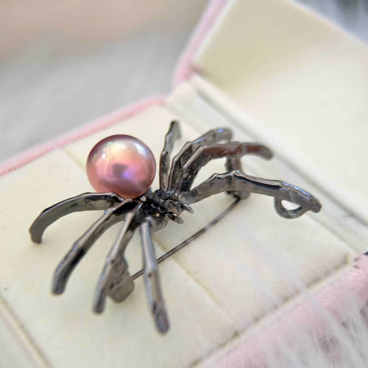 A spider-shaped brooch with a glossy pink pearl and dark metallic legs, displayed on a cream-colored jewelry box.
