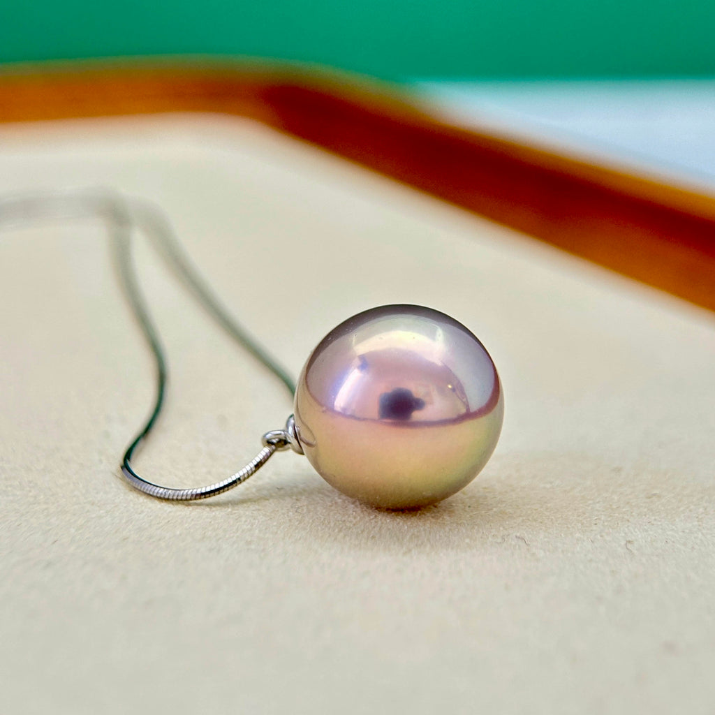 A single round pearl pendant necklace with a silver-toned chain, resting on a light beige textured surface. The pearl has a pearlescent lavender-pink color with a dark reflection in the center.