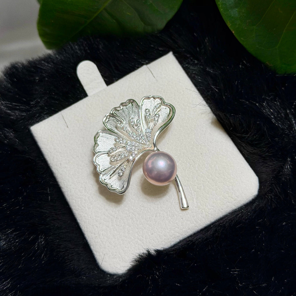 A silver-toned ginkgo leaf brooch with a single pink pearl, displayed on a white jewelry card over a black textured surface with green leaves in the background.