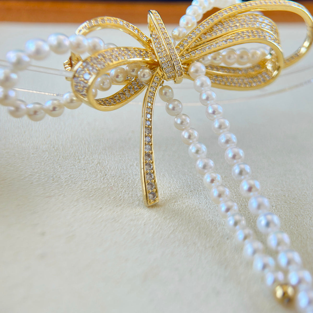 A gold bow-shaped hair claw clip embellished with pavé-set clear stones and adorned with strands of white faux pearls, shown on a light beige textured background.