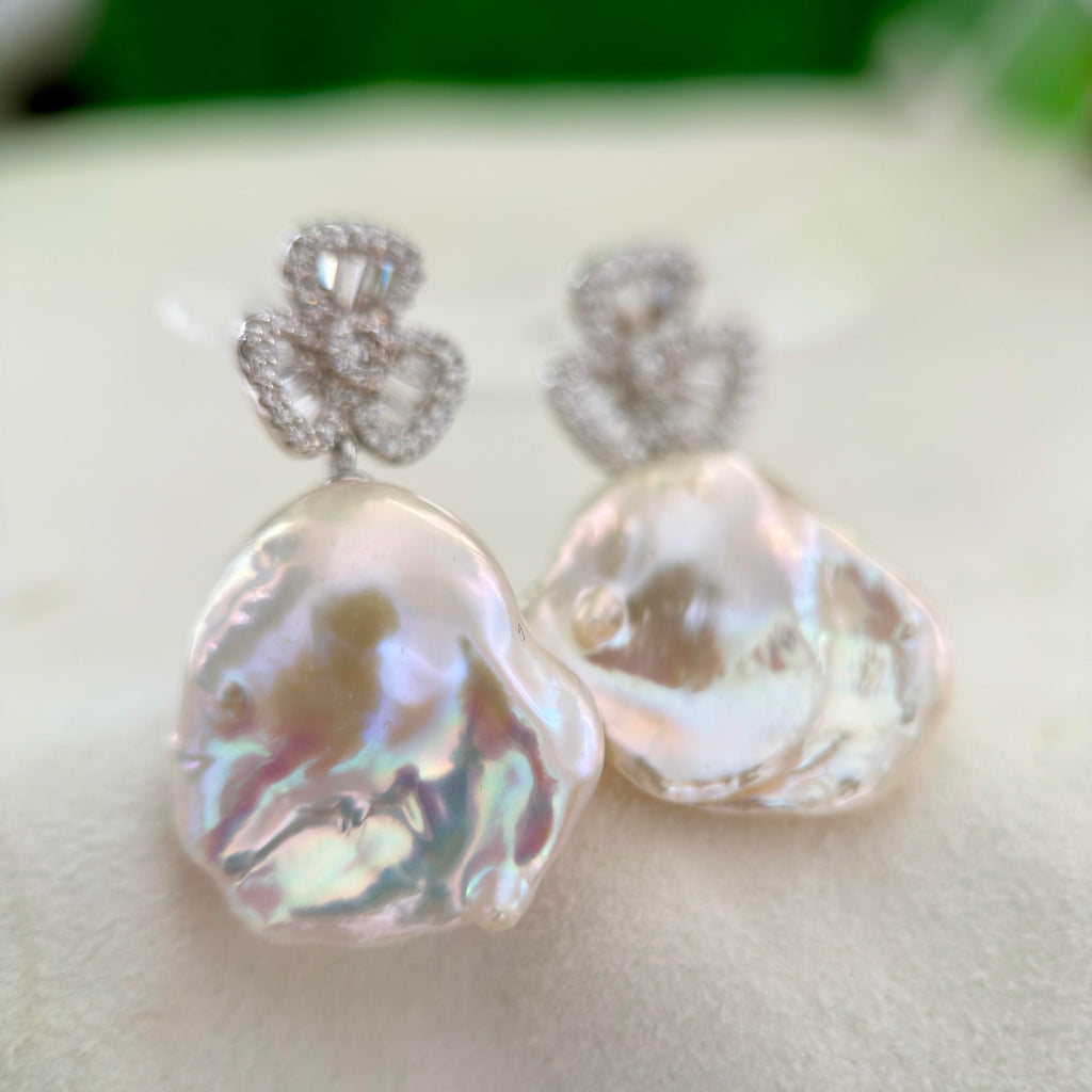 A pair of baroque pearl drop earrings with silver floral studs, displayed on a soft white surface.
