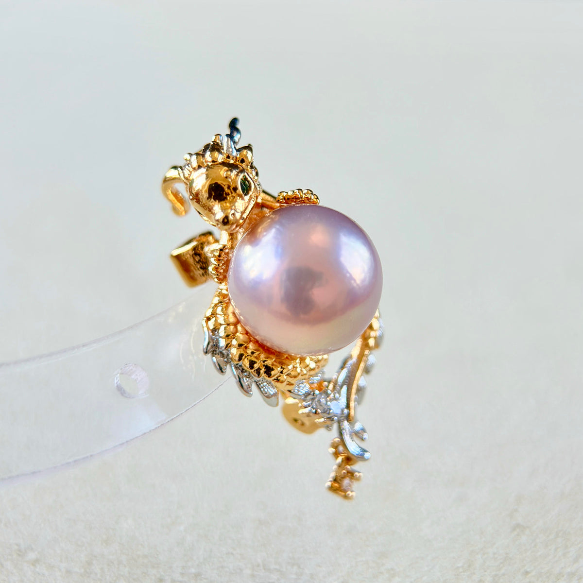 A gold and silver dragon-themed ring featuring a large pink pearl, displayed on a clear plastic jewelry stand against a light beige background.