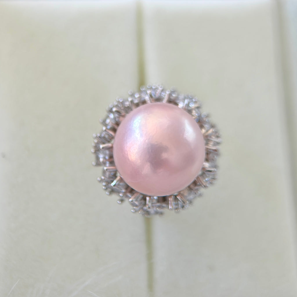 A single ring featuring a round, lustrous light pink pearl surrounded by a halo of small clear stones in a silver-toned setting, displayed on a cream-colored ring box.