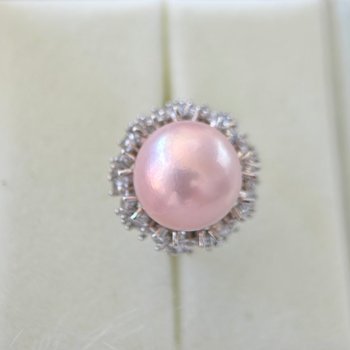A single ring featuring a round, lustrous light pink pearl surrounded by a halo of small clear stones in a silver-toned setting, displayed on a cream-colored ring box.