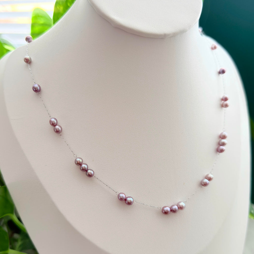 A delicate silver chain necklace with small, irregular pinkish-purple freshwater pearls, displayed on a white mannequin bust with green foliage in the background.