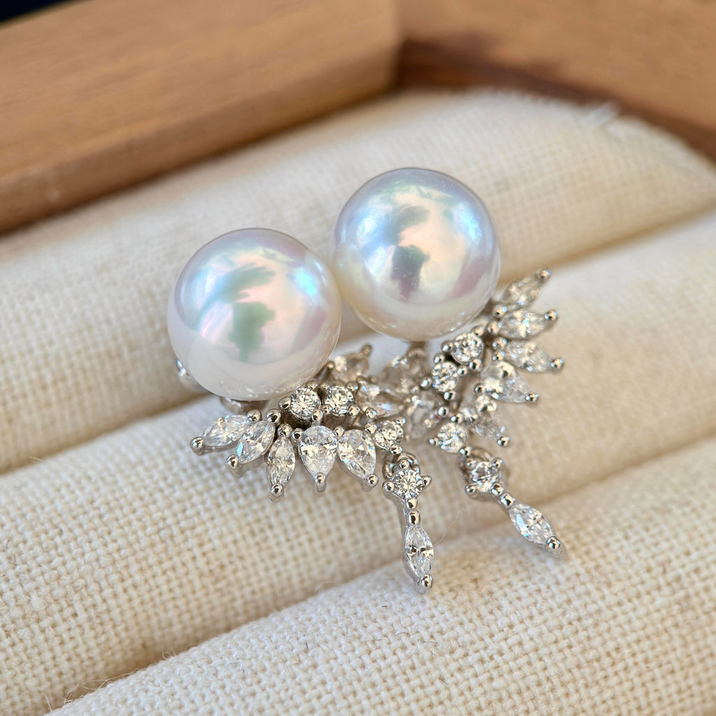 A pair of pearl stud earrings with silver floral leaf settings, resting on a cream-colored textured jewelry display cushion.