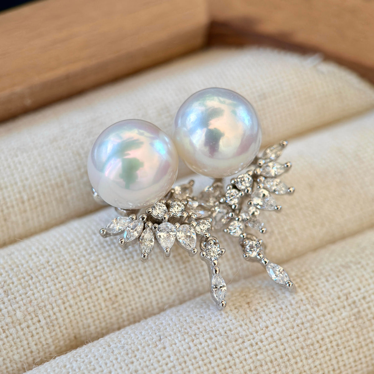A pair of pearl stud earrings with silver floral leaf settings, resting on a cream-colored textured jewelry display cushion.
