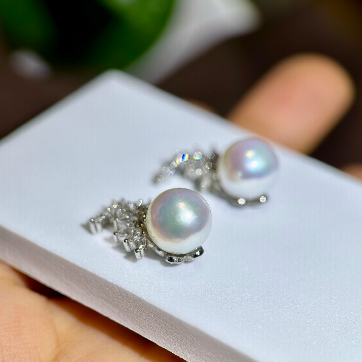 A pair of silver-toned drop earrings with large iridescent white pearls and small clear stones, displayed on a white jewelry card held in a hand.