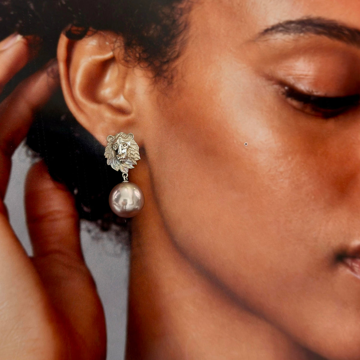 A close-up of a model wearing a silver dangle earring featuring a lion head charm and a single pearl drop.