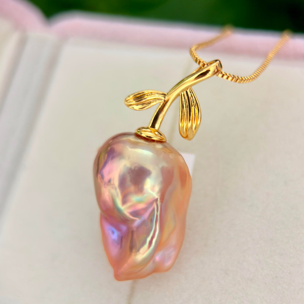 A gold pendant necklace featuring a large, baroque-shaped iridescent pink pearl with a gold leaf-stemmed top, resting on a white jewelry box.