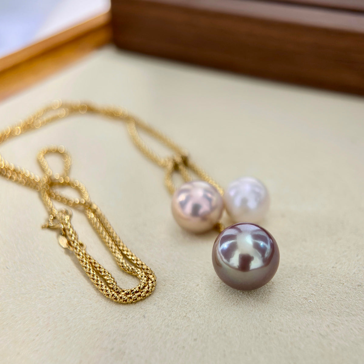 A gold chain necklace with three pearl-like drops in shades of pink, white, and dark gray, displayed on a beige surface.