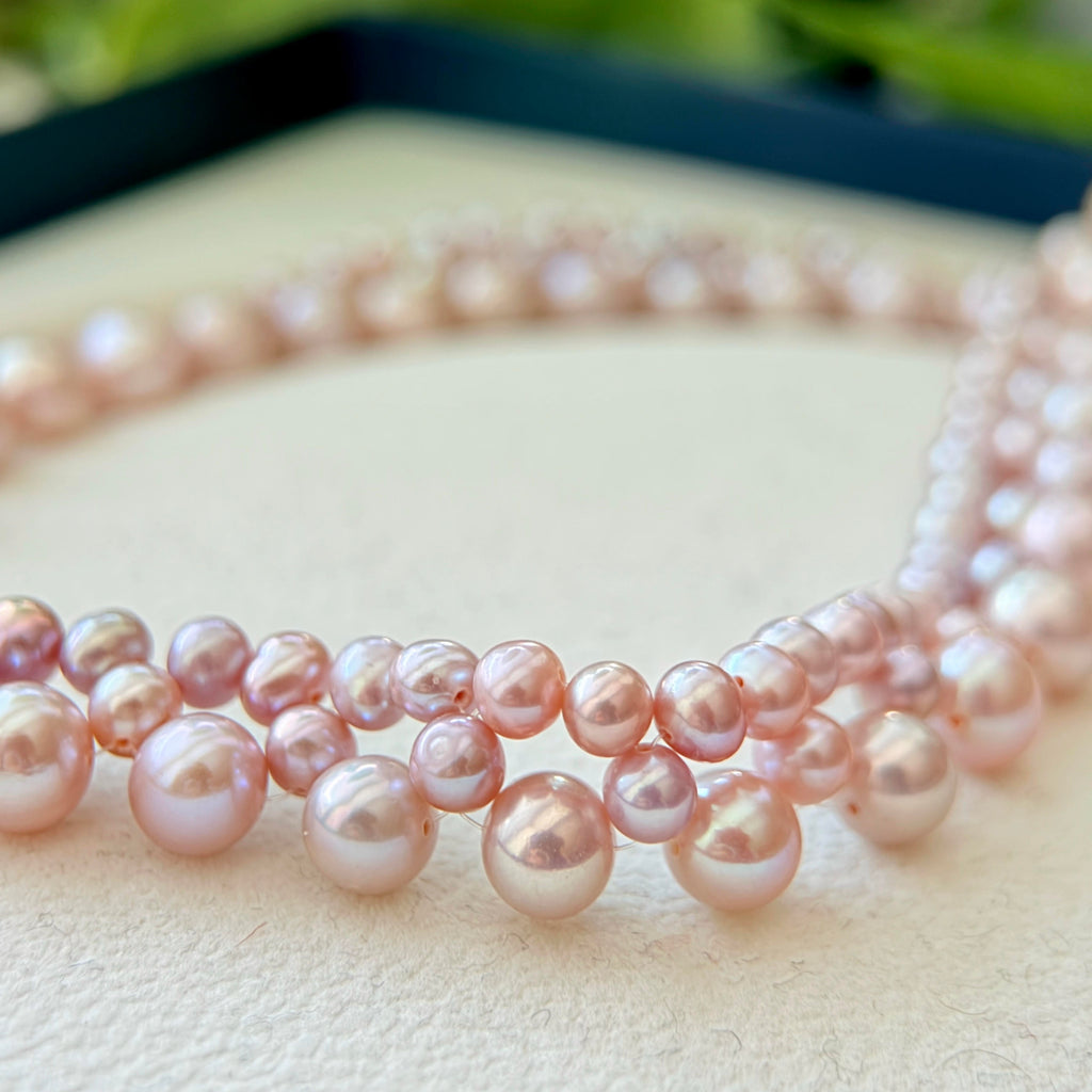 A close-up of a strand of freshwater pearl beads in a soft pink color, arranged in multiple parallel rows on a textured cream surface.
