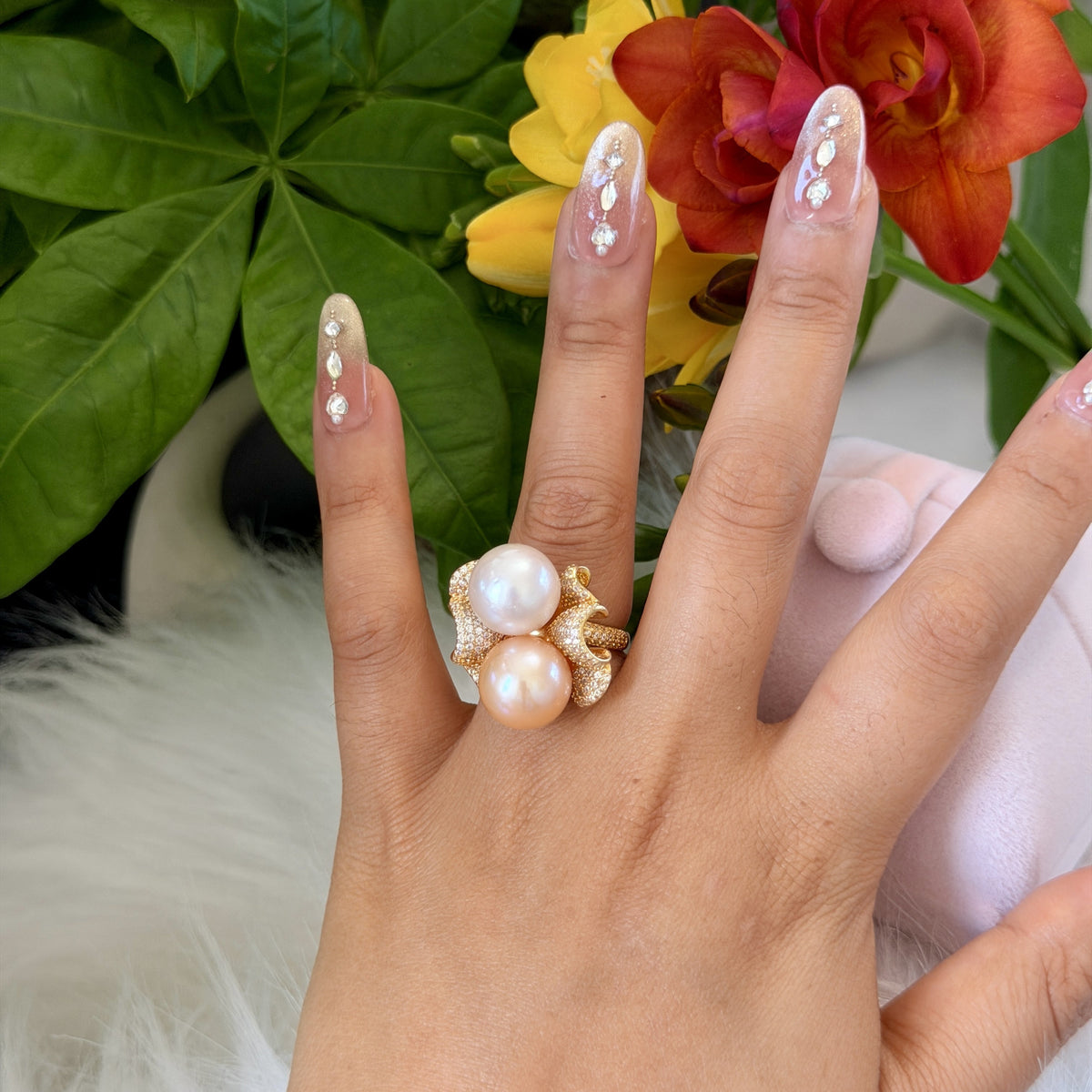 A close-up of a hand wearing a statement ring with two large pearls and gold-toned floral details, set against a background of green leaves and orange flowers.