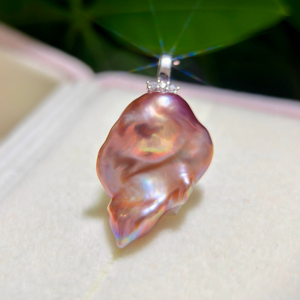 A close-up of a baroque pearl pendant with a silver bezel set with small clear stones, resting on a light cream jewelry box.