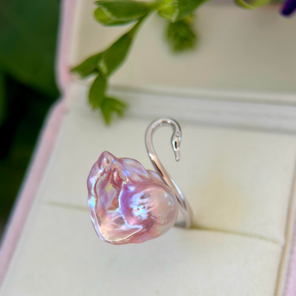 A silver ring shaped like a swan, featuring a large, organic-shaped pink iridescent pearl on its body, displayed inside an open cream-colored jewelry box with blurred green leaves in the background.