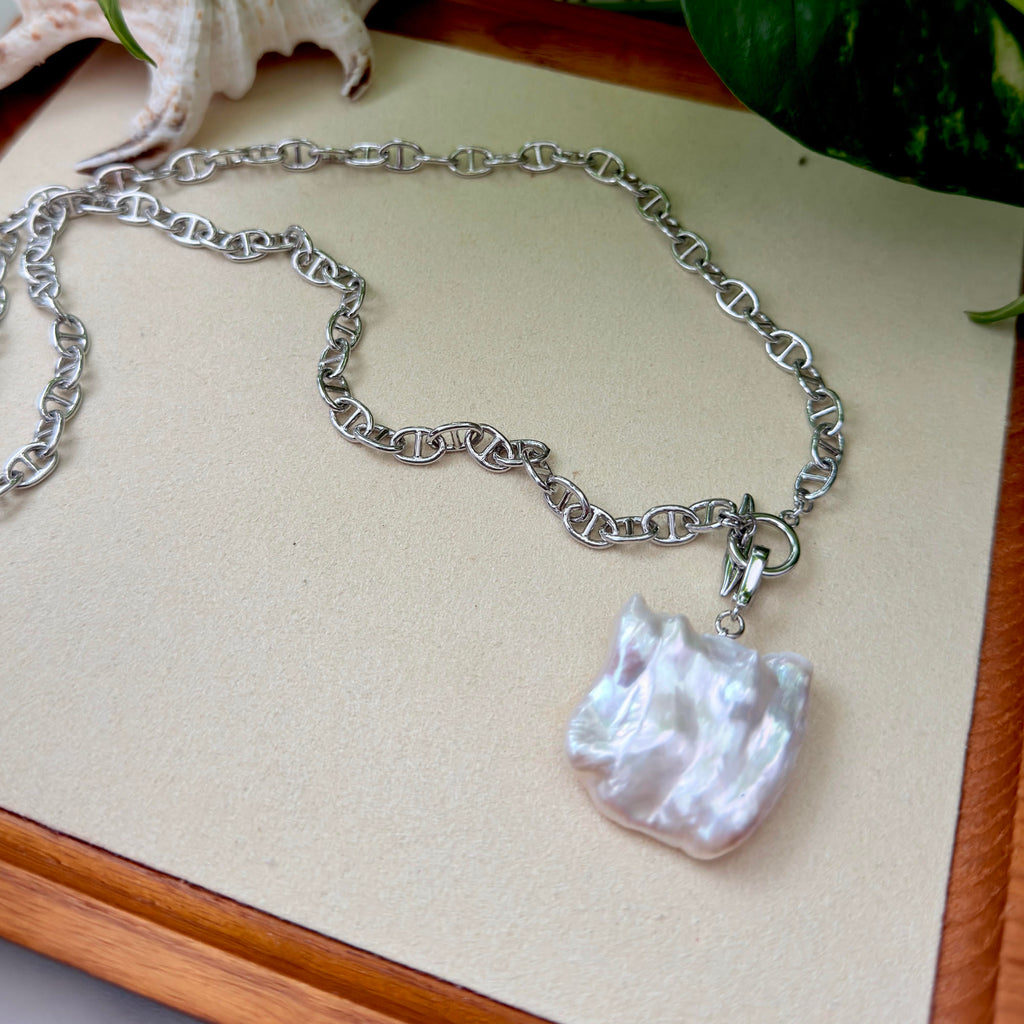 A silver chain necklace with a large, irregular white baroque pearl pendant, displayed on a beige textured card with a wooden border.