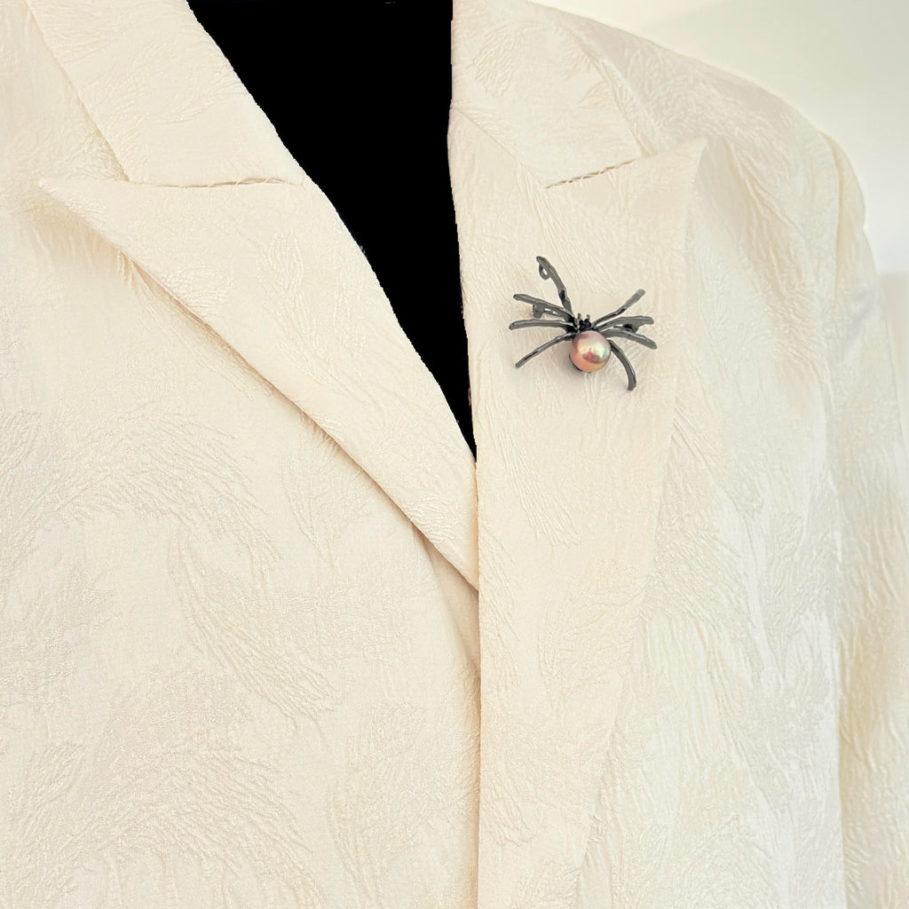 A close-up of a textured cream-colored blazer with a small spider brooch pinned to the chest. The spider brooch features a black body and legs with a small pink pearl at its center, set against a black mannequin torso.