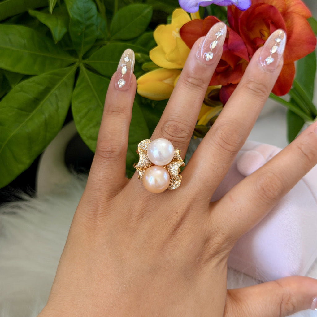 A close-up of a hand wearing a gold ring with two large white pearls, set against a background of colorful flowers and green leaves.