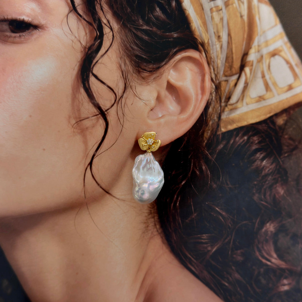 A close-up of a person wearing a gold post earring with a large, irregular white baroque pearl drop, with dark curly hair and a patterned headscarf visible.