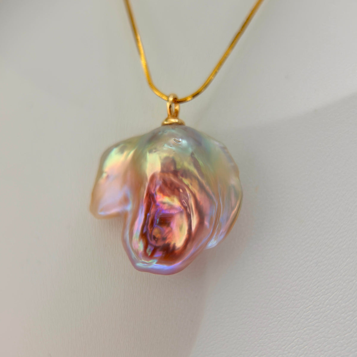 A gold necklace featuring an organic, baroque-shaped iridescent pearl pendant with shimmering pink, green, and blue hues, displayed against a clean, light off-white background.