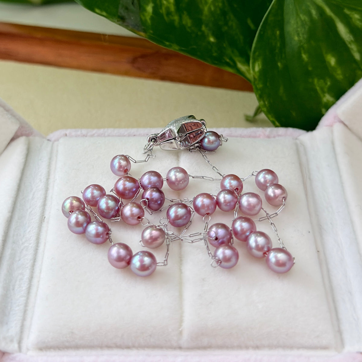 Purple freshwater pearl drop earrings with silver chain links, displayed on a light cream surface with a soft-focus green leaf background.