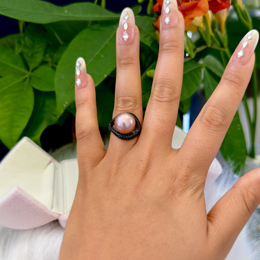 A close-up of a hand wearing a ring with a large pink pearl set in a textured black band, with decorative nail art and a floral background.