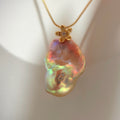 A gold flower-shaped pendant necklace with a baroque-style iridescent pearl pendant, displayed on a cream-colored jewelry stand.