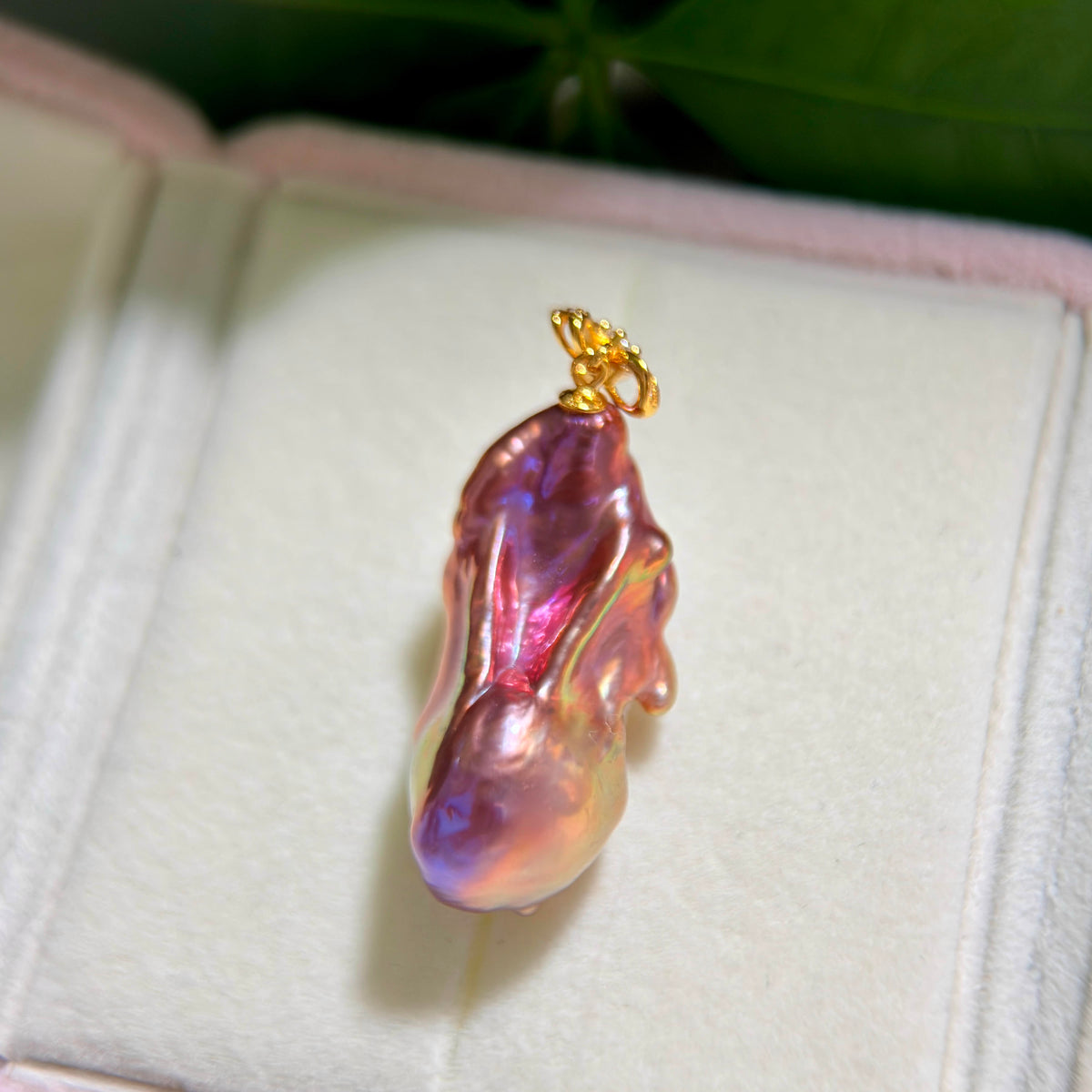 A baroque pearl pendant with iridescent pink, purple, and blue hues, set in a gold bail, displayed on a white jewelry box.
