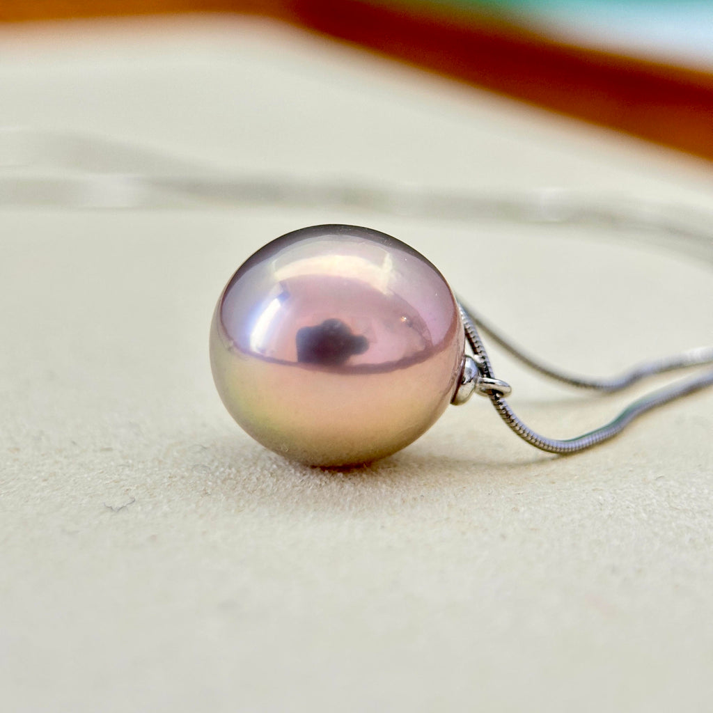 A single round pearl pendant necklace with a silver-toned chain, resting on a light beige textured surface.