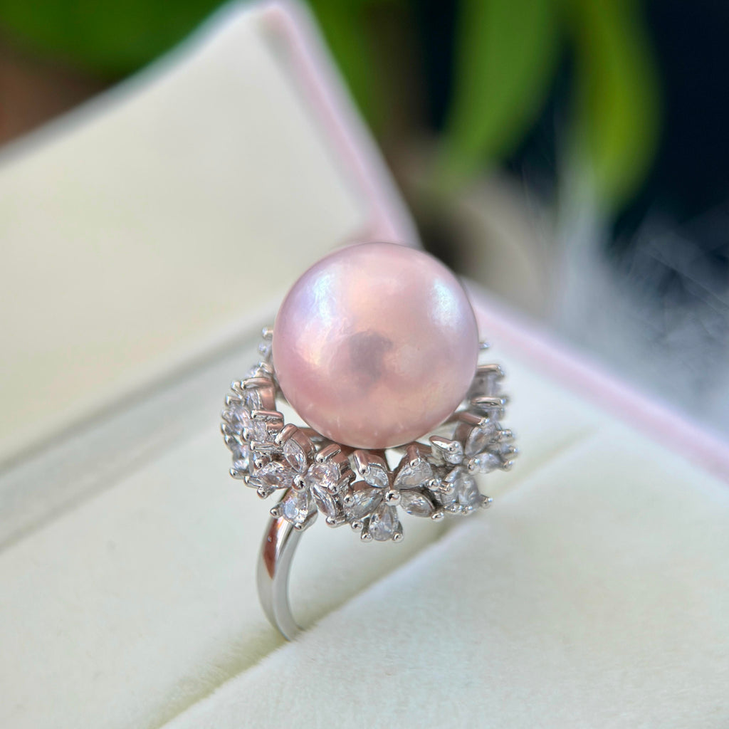 A pink freshwater pearl ring with a silver-toned floral halo and clear accent stones, displayed on a cream-colored jewelry box.