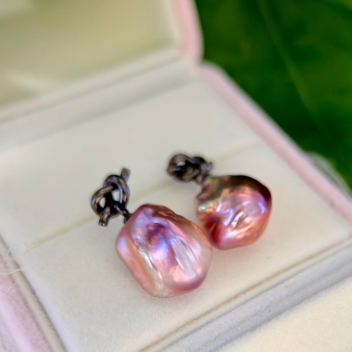 A pair of baroque pearl stud earrings with iridescent pink and purple pearls on dark metal posts, displayed inside an open light pink jewelry box.