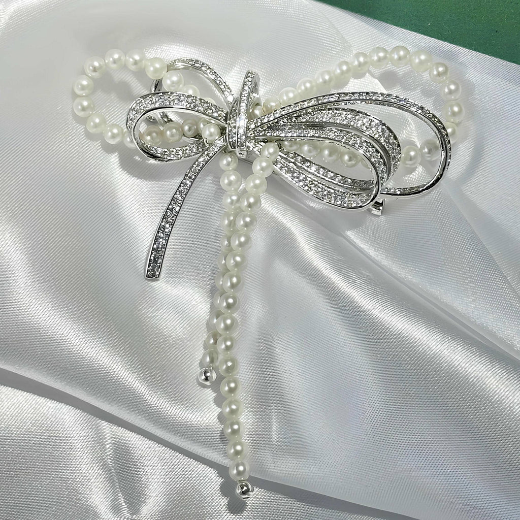 A silver bow-shaped brooch decorated with white faux pearls and sparkling clear crystals, displayed on a white satin fabric background.