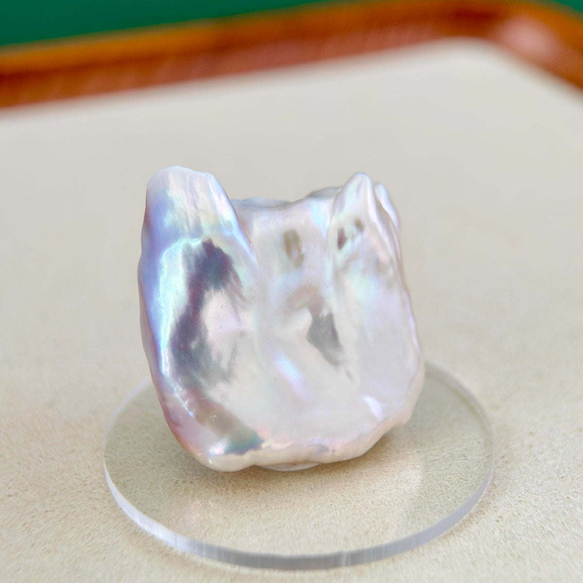 A single irregular baroque pearl with a pearly white luster and iridescent rainbow reflections, displayed on a clear round acrylic stand against a neutral beige background.