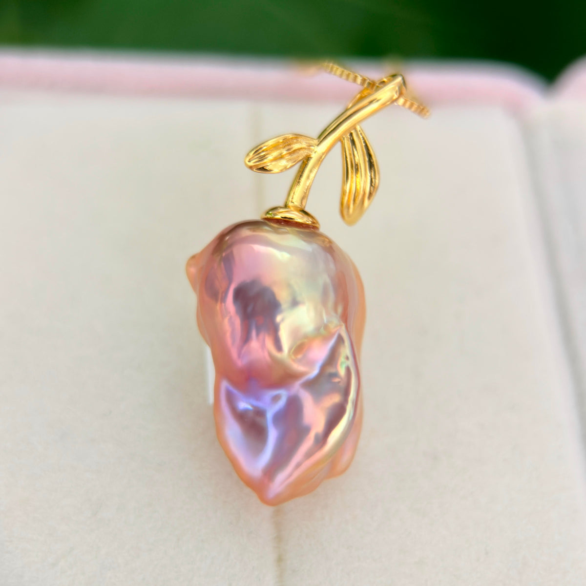 A close-up of a gold pendant shaped like a pearl fruit, resting on a light cream jewelry box.