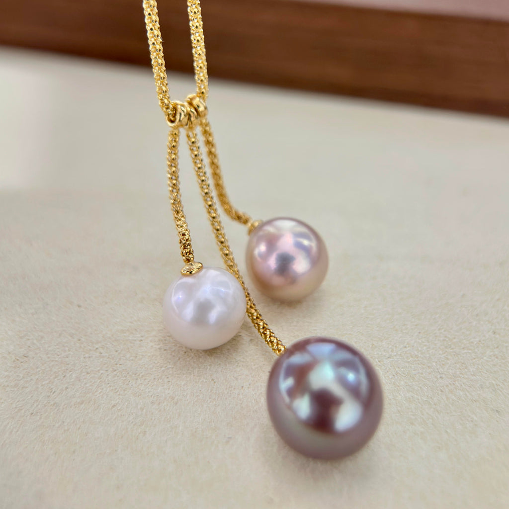 A close-up of a gold chain necklace with three dangling pearl-like beads in white, pale pink, and a larger iridescent lavender-gray pearl, resting on a light beige surface.