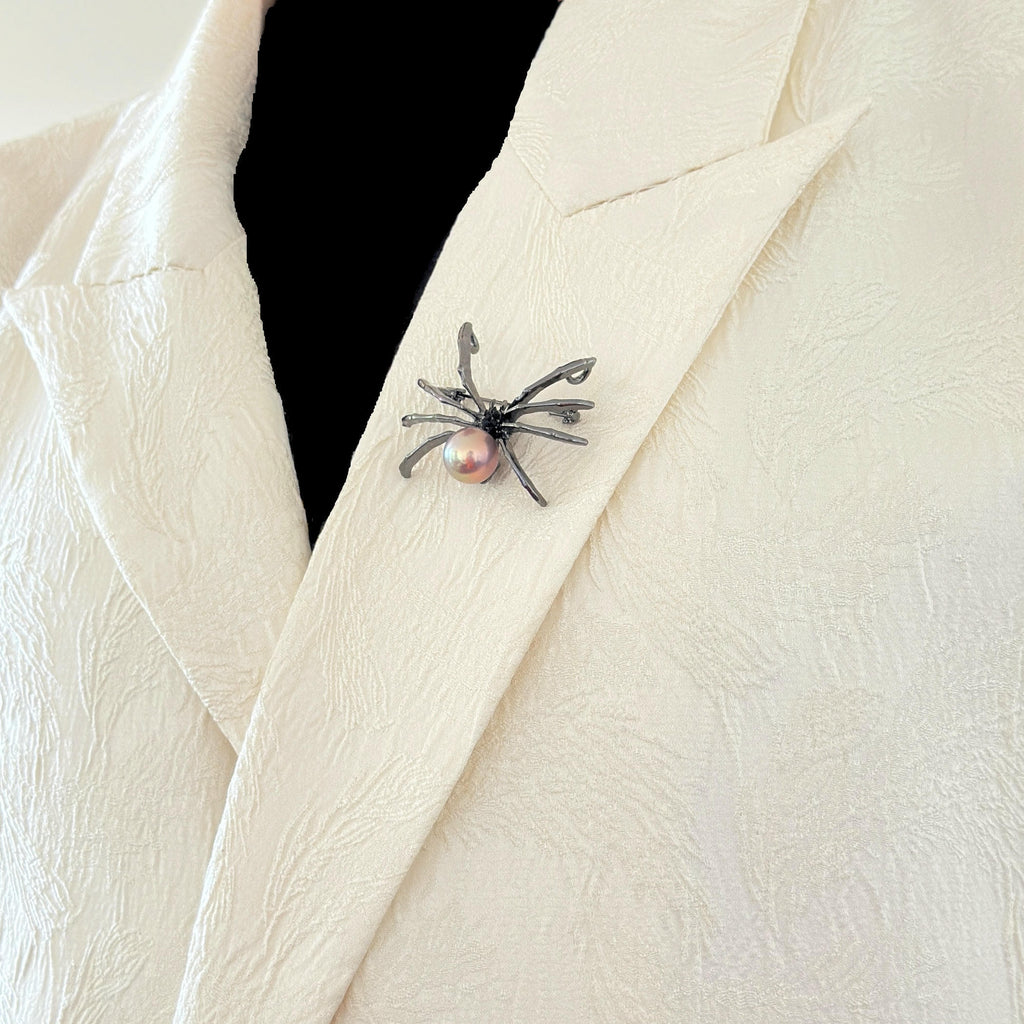 A close-up of a textured cream-colored jacket with a spider-shaped brooch pinned to the lapel. The brooch features a dark metallic spider body with a small, round pink pearl at its center.