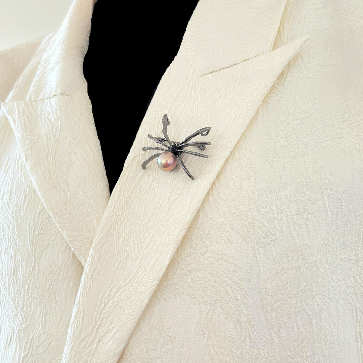 A close-up of a textured cream-colored jacket with a spider-shaped brooch pinned to the lapel. The brooch features a dark metallic spider body with a small, round pink pearl at its center.
