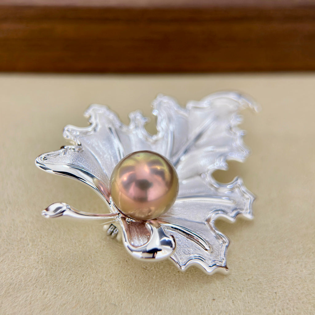 A silver-toned leaf-shaped brooch with a central round pearl in a warm pinkish-gold tone, photographed close up on a beige surface.