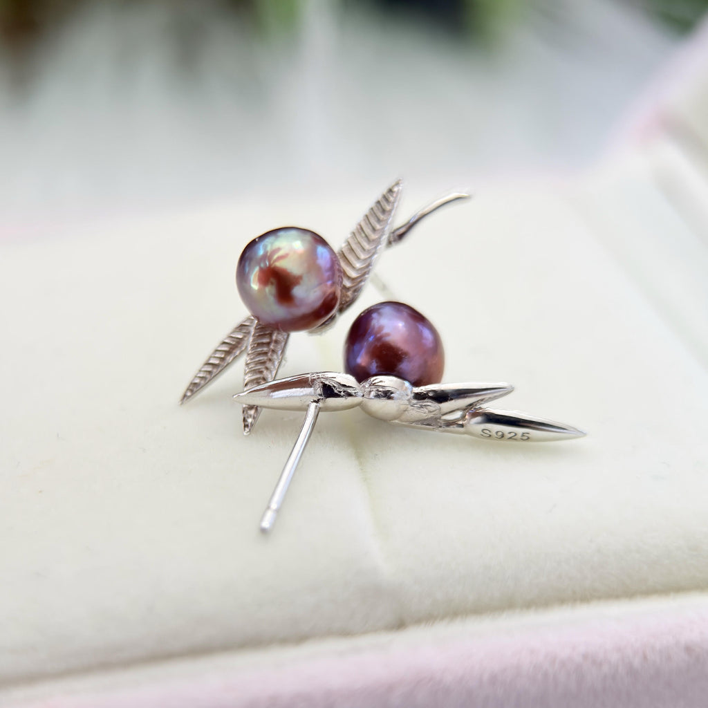 A pair of silver dragonfly stud earrings with iridescent purple pearls, displayed on a soft cream-colored jewelry box.